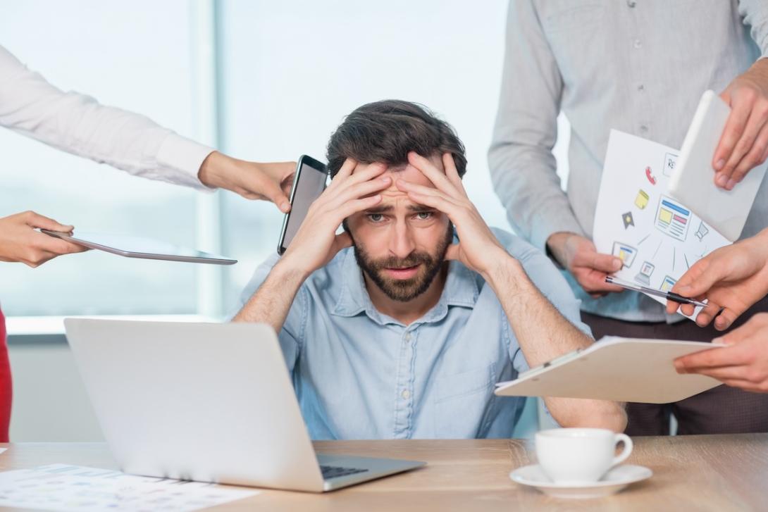 Frustrated man surrounded by technology, papers, people.